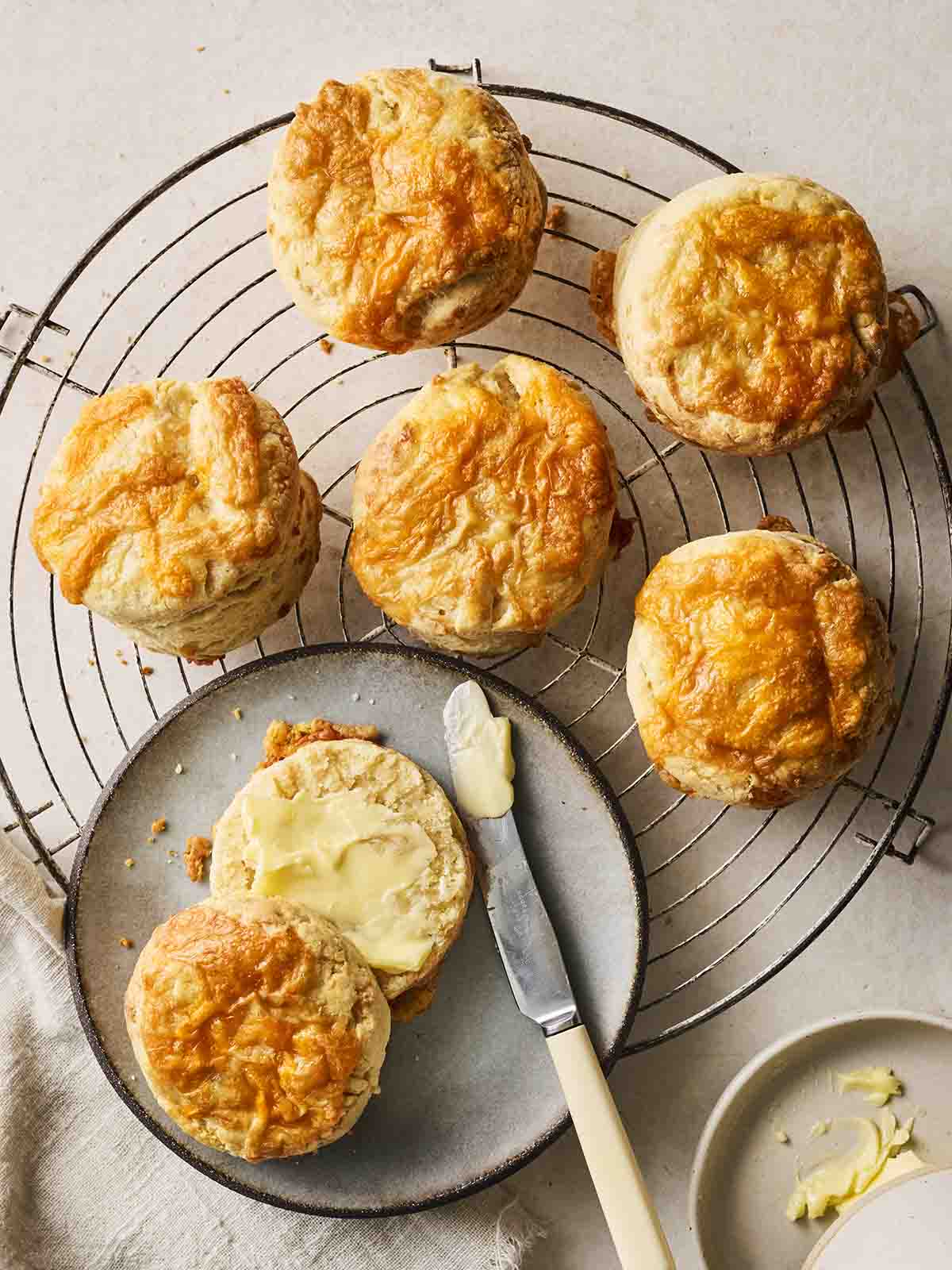 A round wire rack with baked Cheese Scones on and one buttered on a plate.