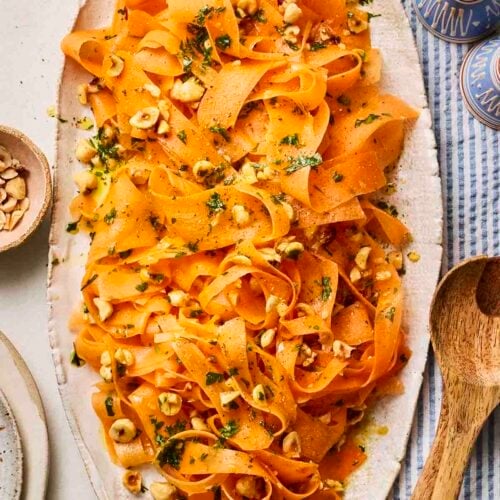A platter of carrot salad with hazelnuts on a table with wooden utensils, salt and pepper and a blue and white cloth.