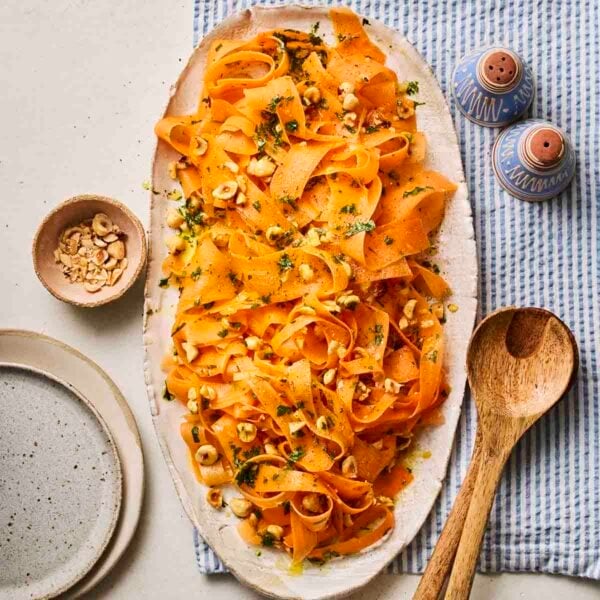 A white platter serving Carrot Salad, on a table with wooden salad servers, salt and pepper pots and a small bowl of hazelnuts to the side.