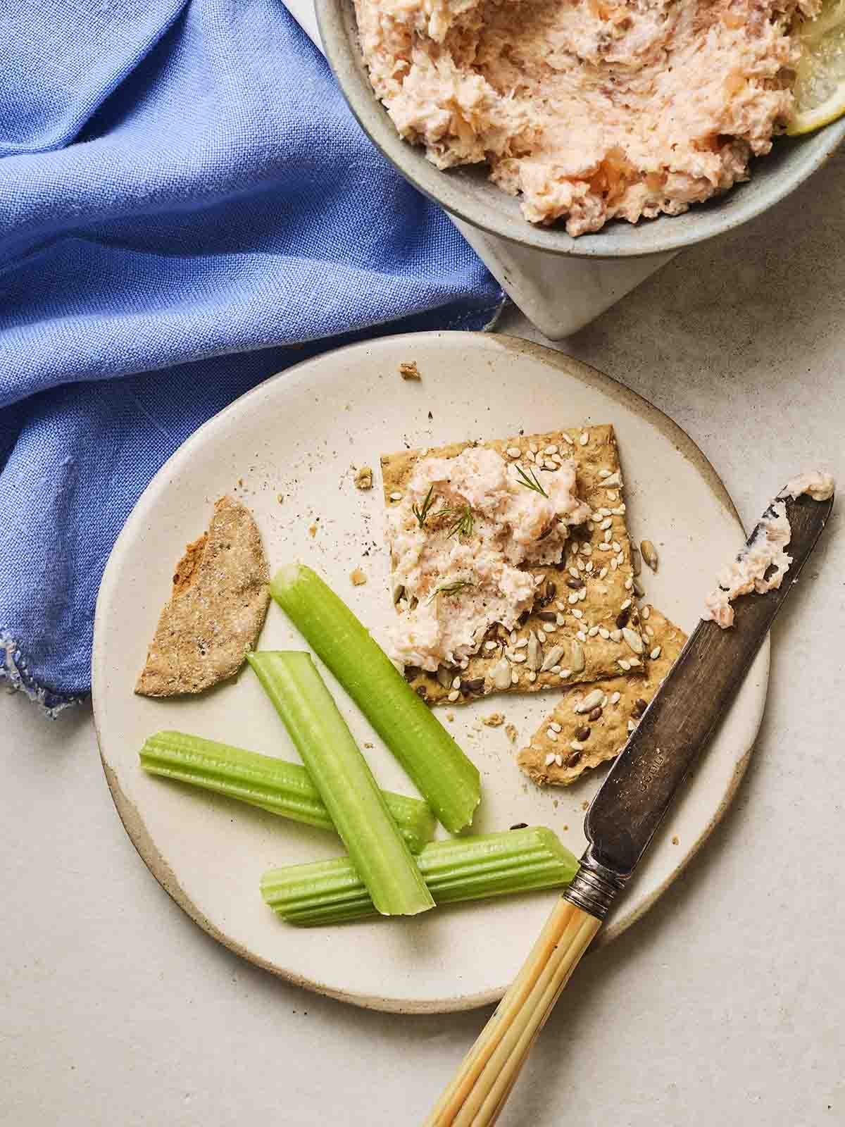 A plate with crackers and smoked salmon pate spread on with a knife, and sticks of celery, with a bowl of pate to the side.