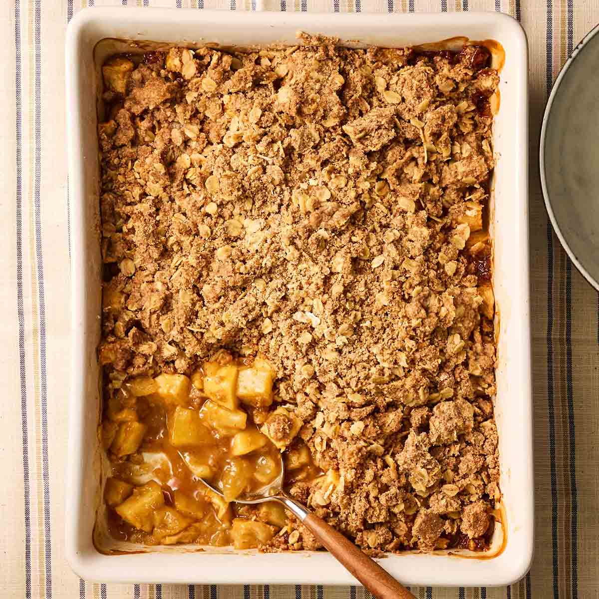 The corner of a pie dish with an Apple Crumble inside, with a spoonful missing, showing the apple filling and golden, crunchy topping, with a spoon sticking out.