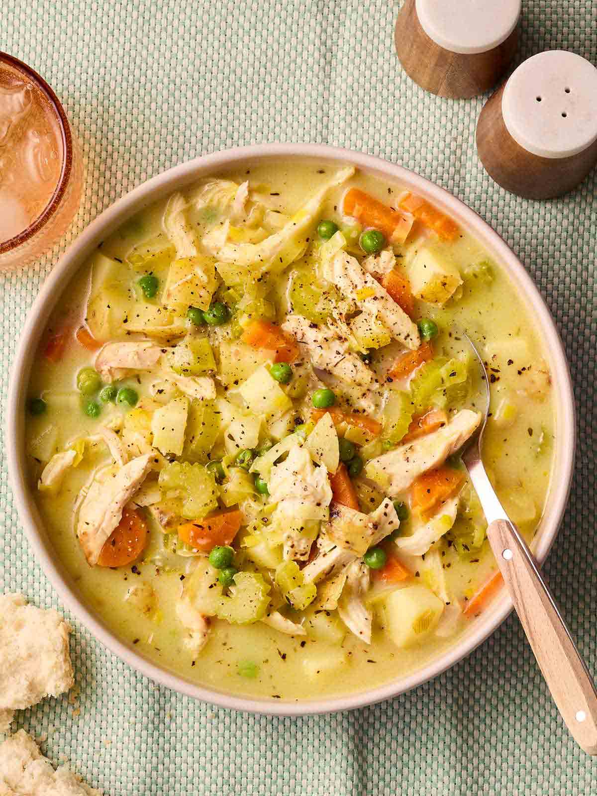 A bowl of chunky chicken soup with a spoon in on a green tablecloth with salt and pepper to the side.