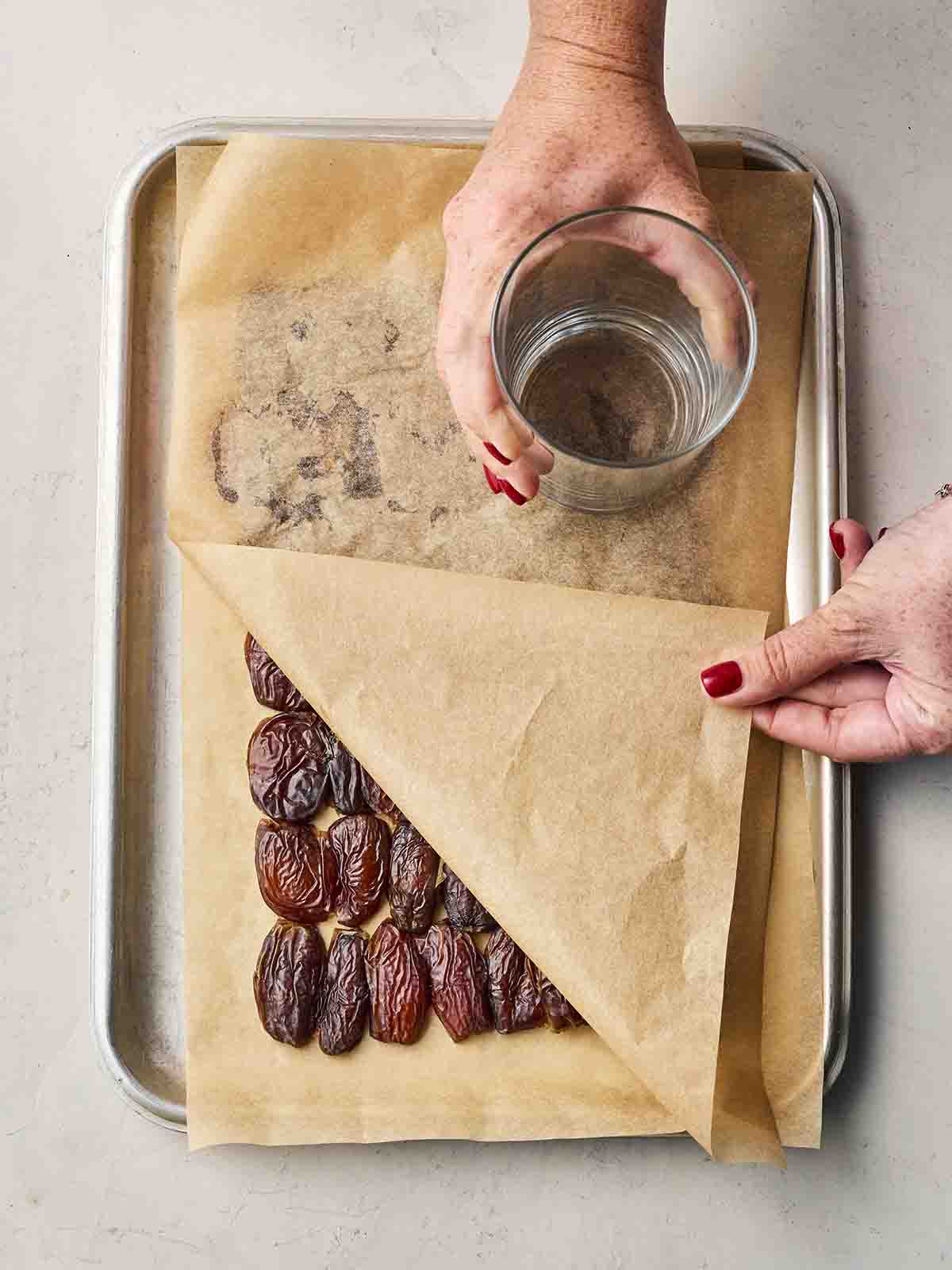 Rows of dates on a baking tray, covered with baking parchment and a hand using a glass to squash them down.