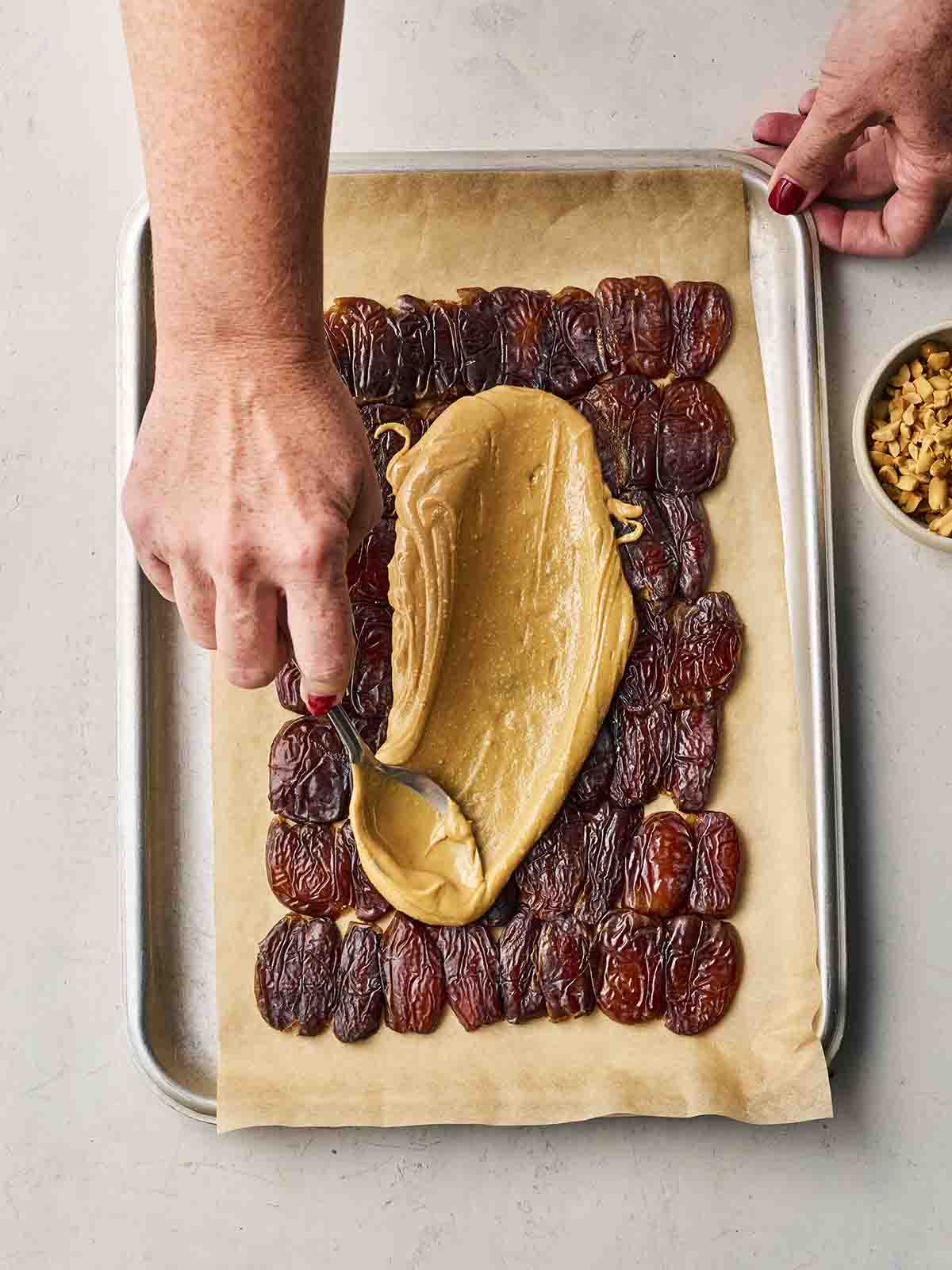 Rows of dates on a tray with two hands spreading over peanut butter.