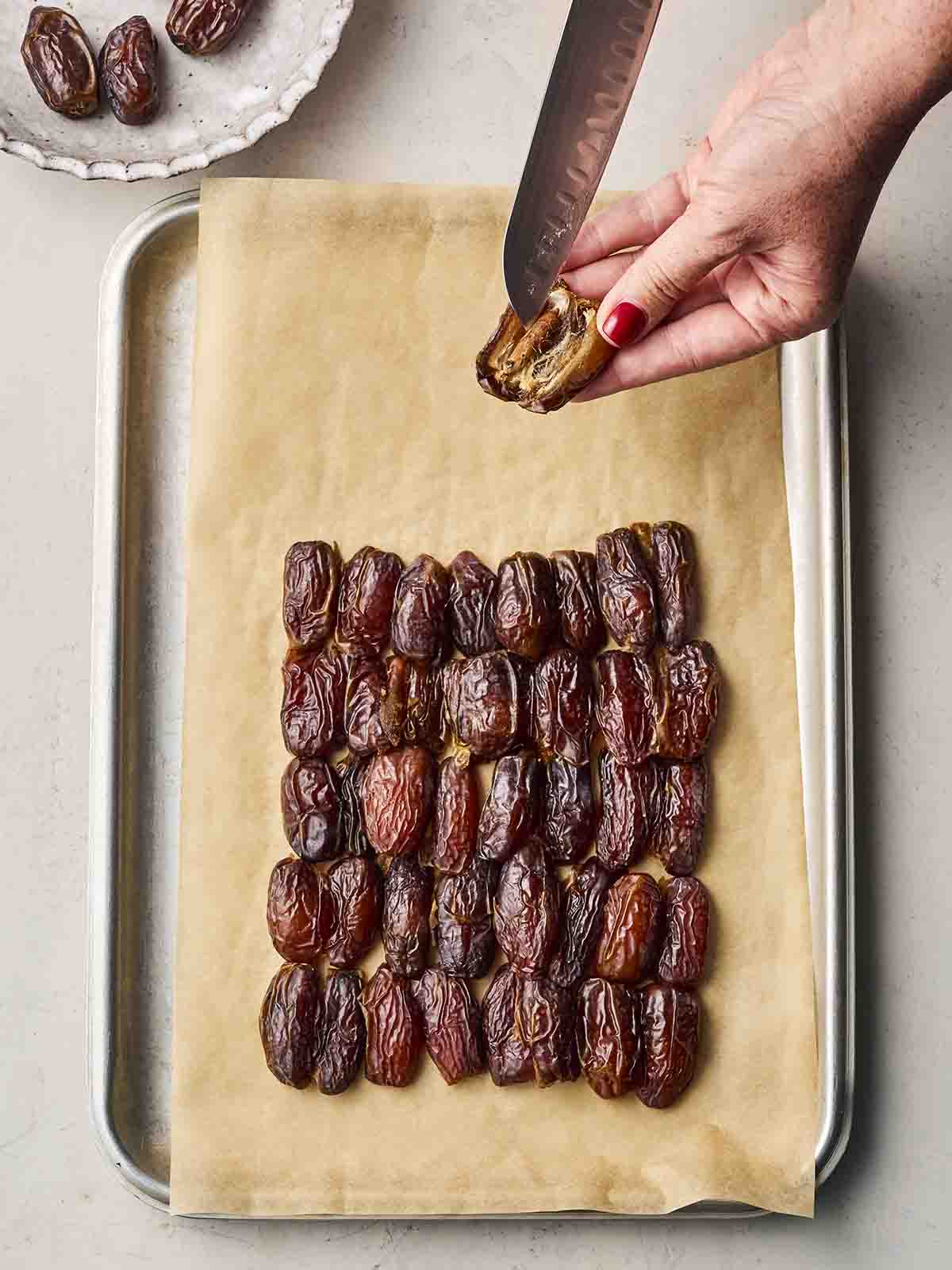A baking tray with rows of dates, with two hands holding on and slicing.
