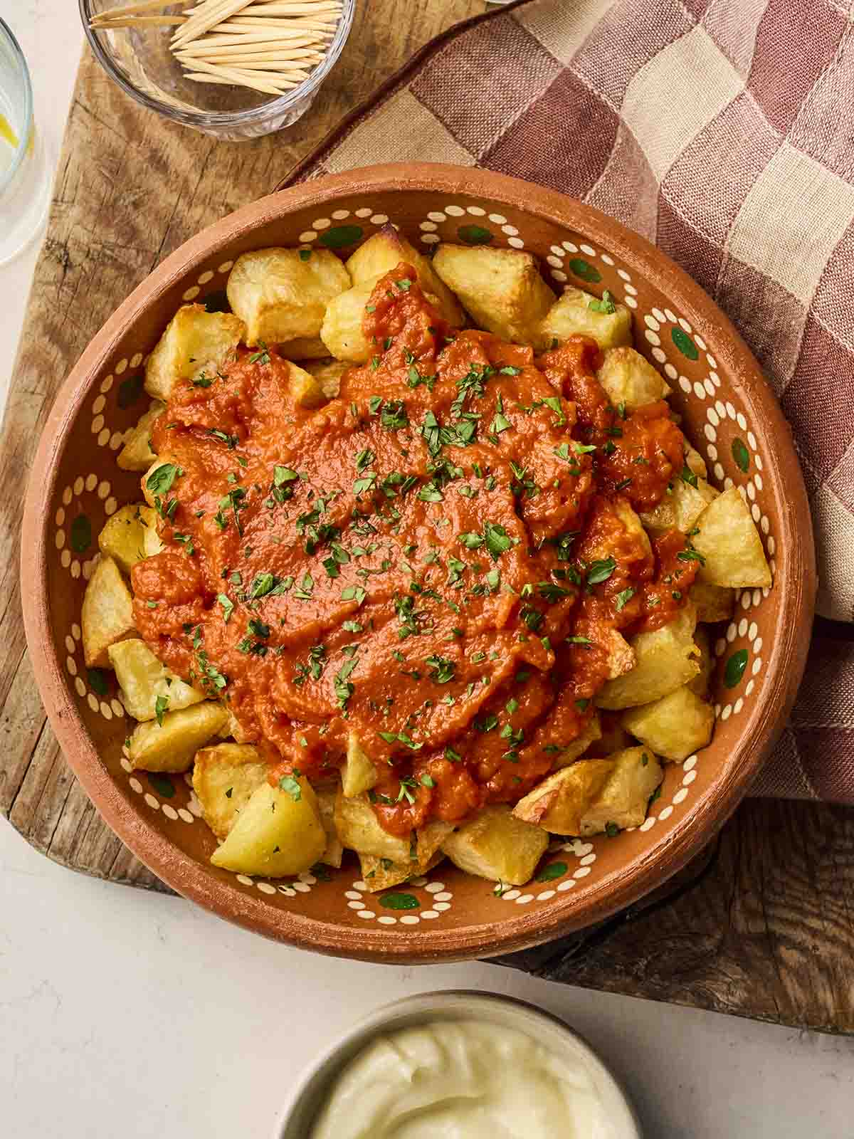 A clay dish on a board filled with Patatas Bravas, ready to eat.