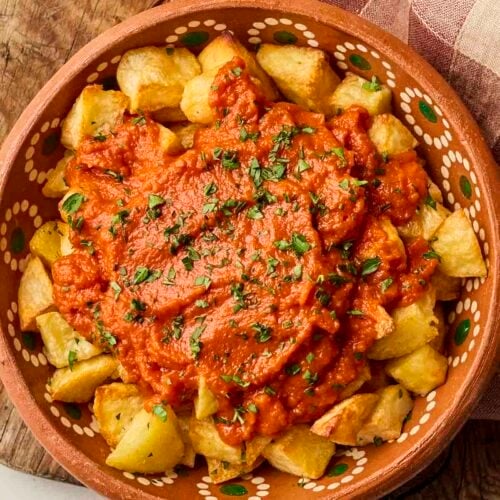 A bowl of Patatas Bravas tapas on a board with a tea towel.