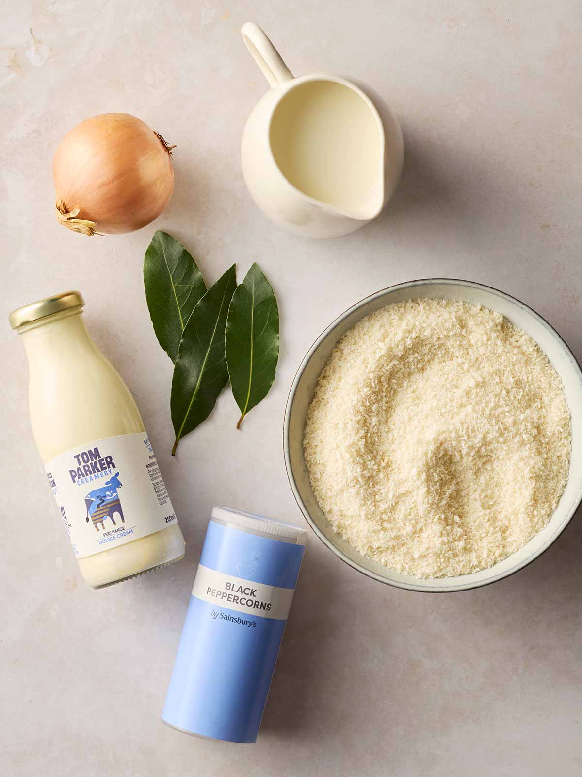 Ingredients for making homemade Bread Sauce laid out on a white counter, including breadcrumbs, milk, bay leaves, onion, black peppercorns and cream.