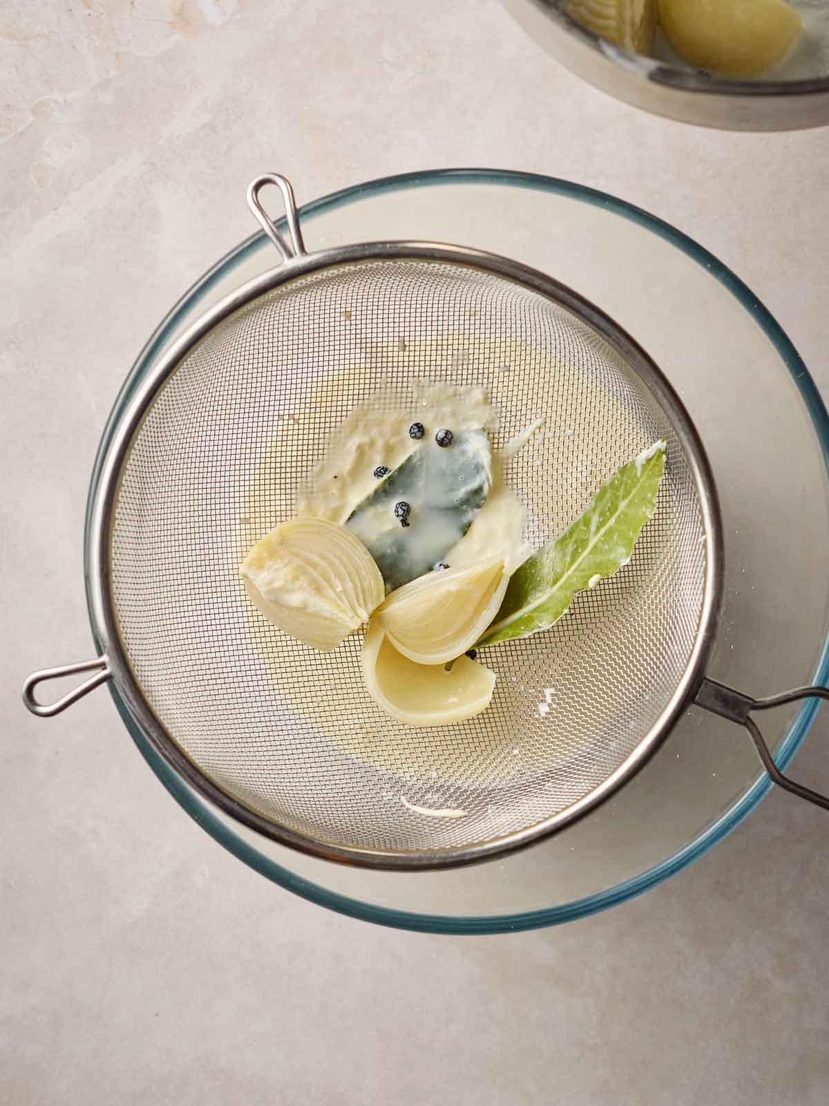 A sieve over a glass bowl, with sliced onion and bay leaves in, with milk in the bowl.