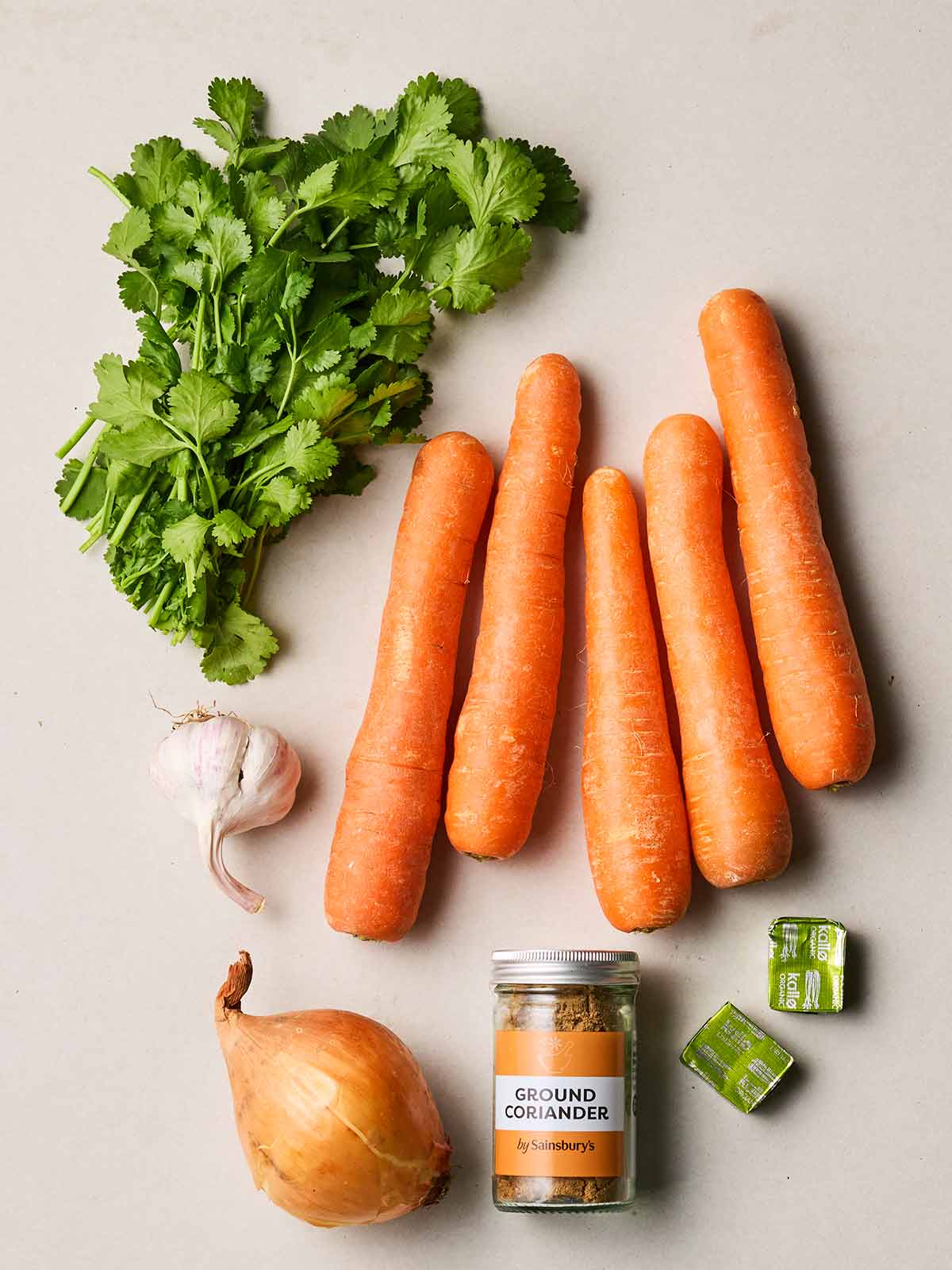 Raw ingredients laid out on a white counter, including carrots, coriander, stock cubes, onion and garlic.