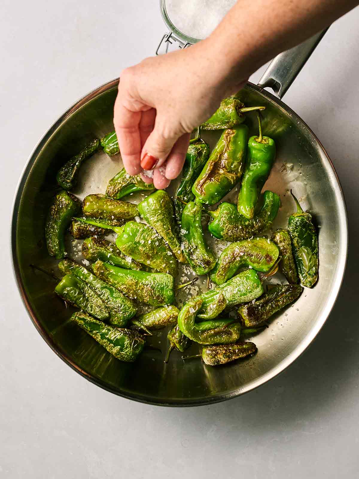 A metal pan filled with charred padron peppers, with a hand sprinkling sea salt over the top.