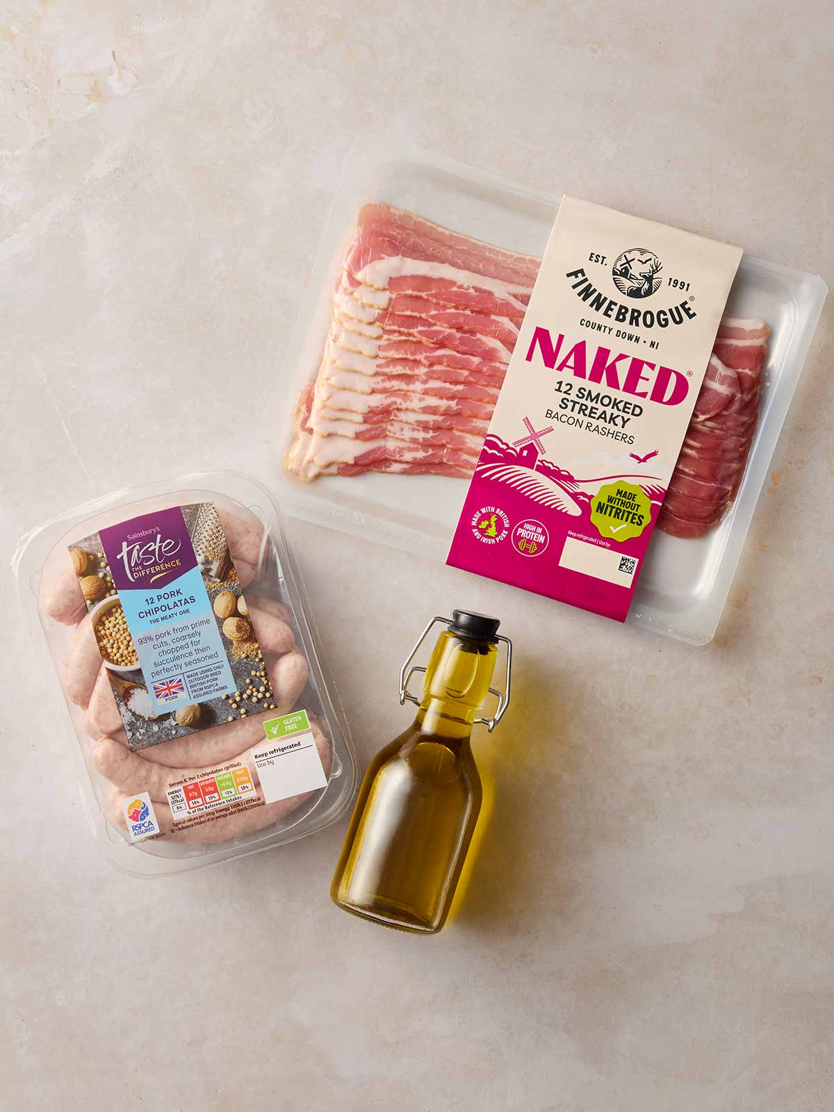 The ingredients for everything you need for making Pigs in Blankets at home laid out on a white surface, including bacon, sausages and oil.