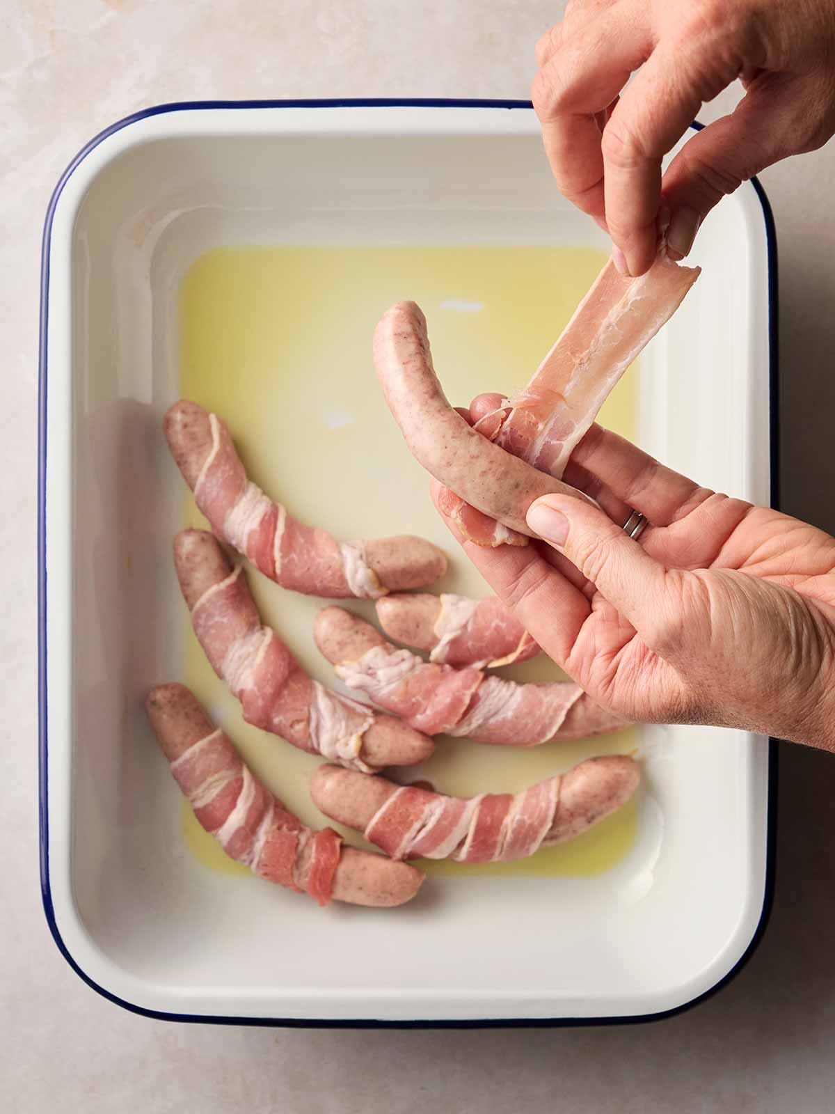 A white oven dish with oil in and sausages wrapped with bacon around, and a hand wrapping up a raw Pig in Blanket.
