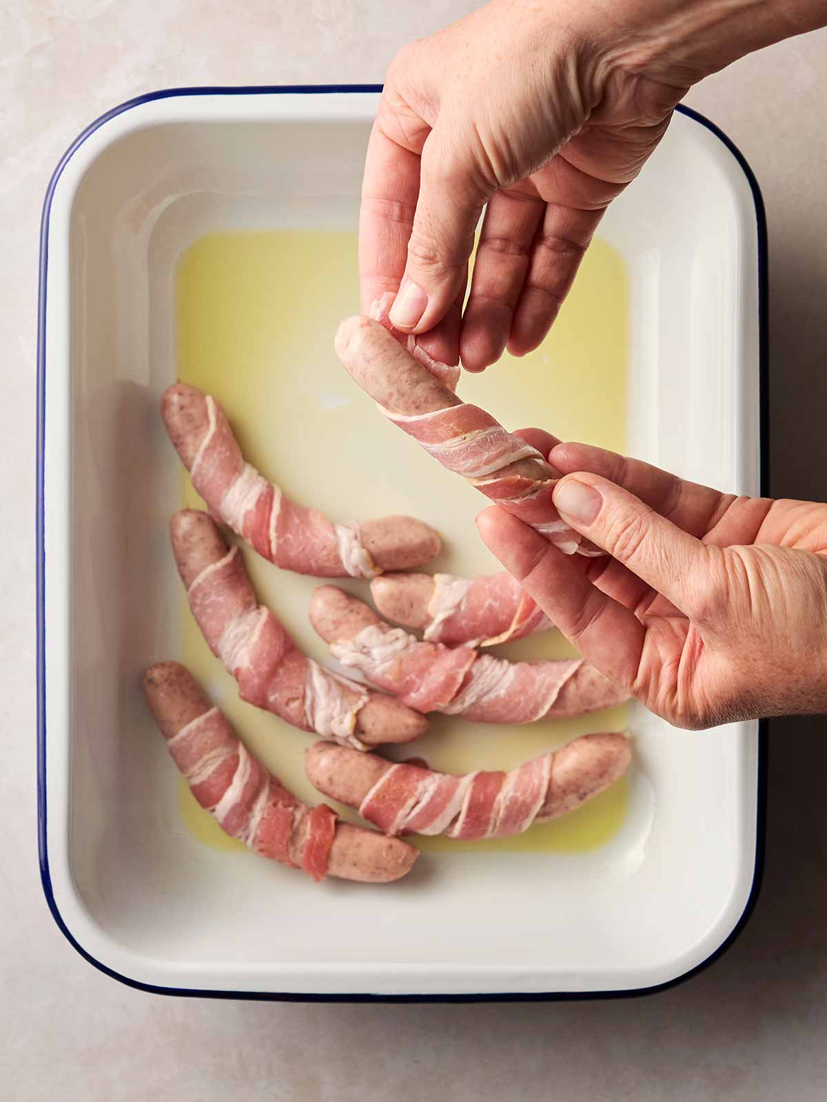 Two hands wrapping bacon around sausages with an oven dish ready to cook Pigs in Blankets.