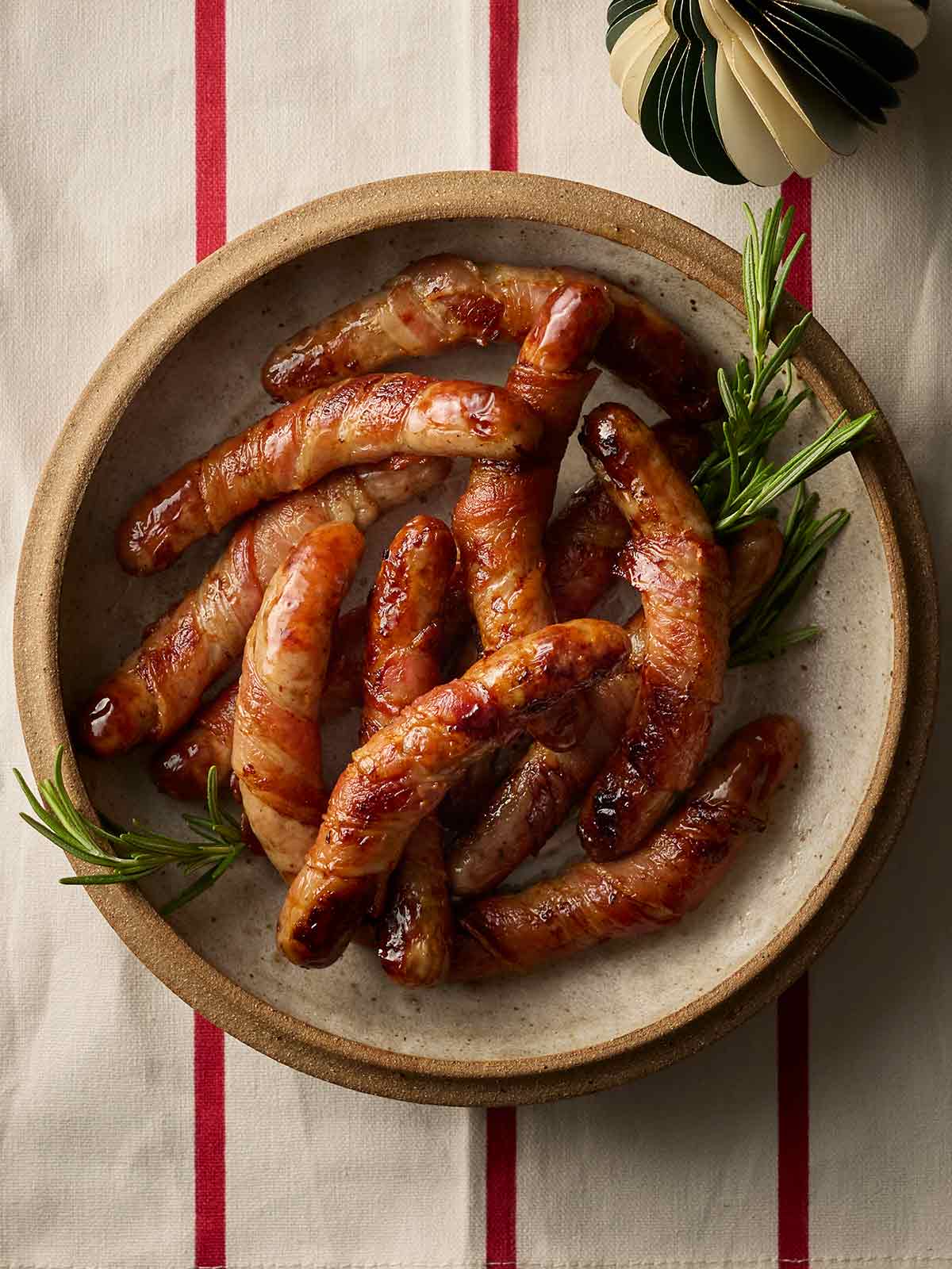 A plate of cooked Pigs in Blankets with sprigs of rosemary, on a red and white stripy tablecloth.