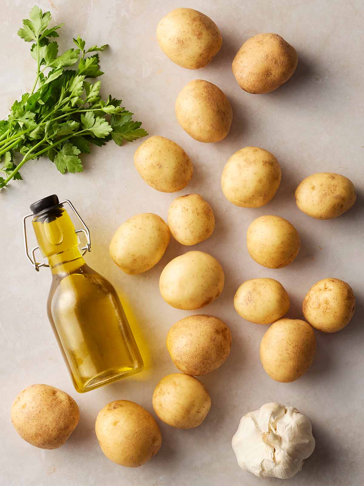 Raw ingredients laid out on a white surface, including potatoes, parsley, oil and garlic.