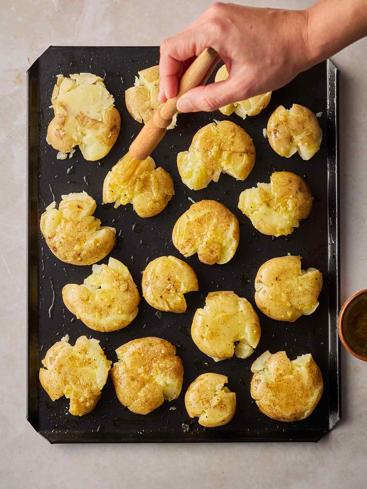 A baking tray with smashed potatoes being brushed with olive oil by a hand.