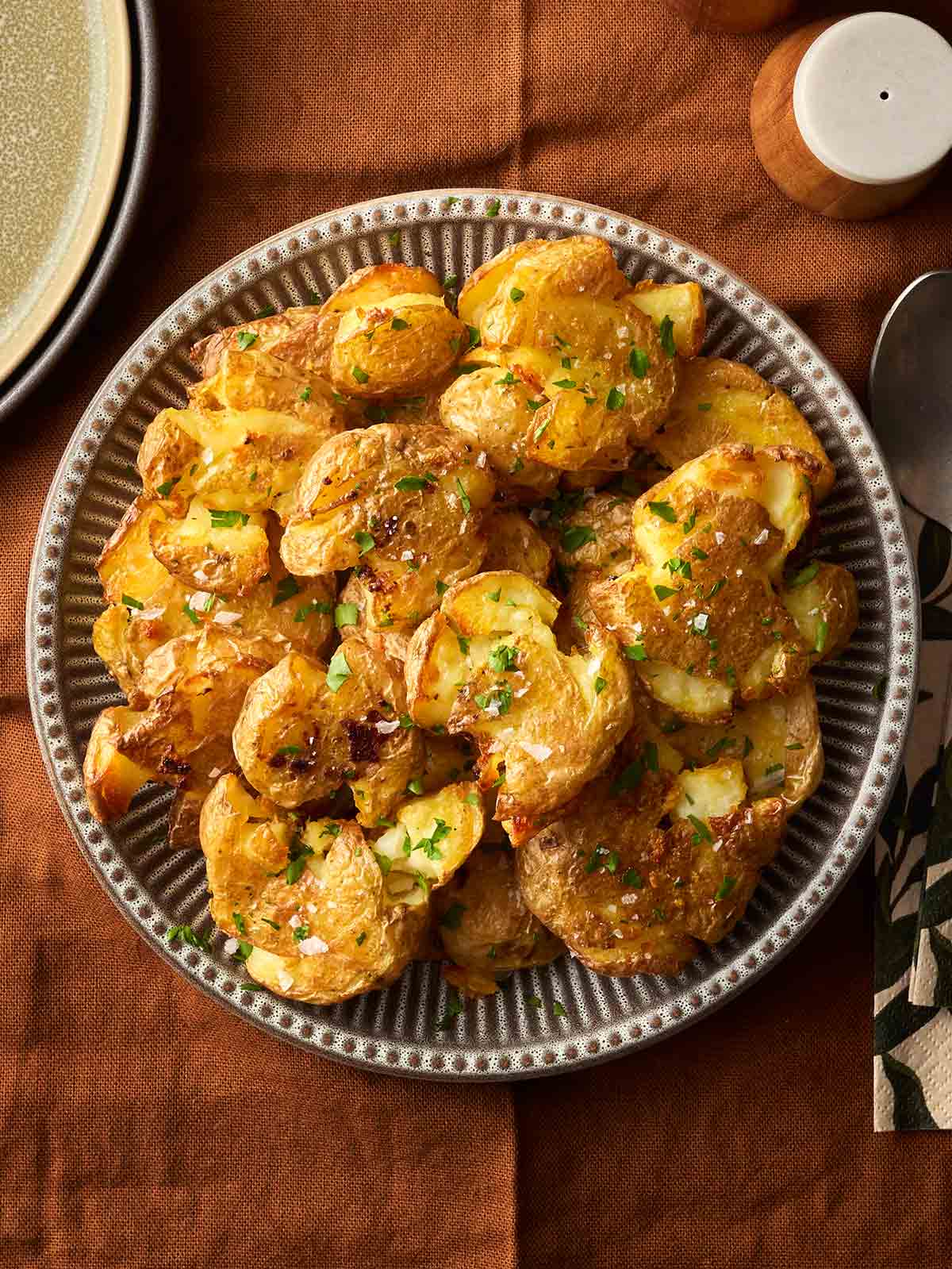 A bowl filled with Smashed Potatoes and garnished with sea salt and parsley on a brown tablecloth.