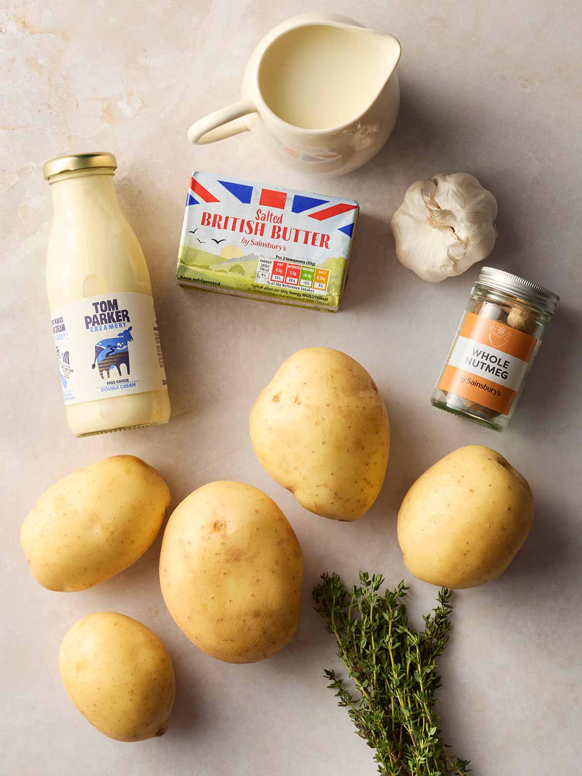 Raw ingredients laid out on a table, including potatoes, cream, butter, nutmeg, milk, thyme and garlic.