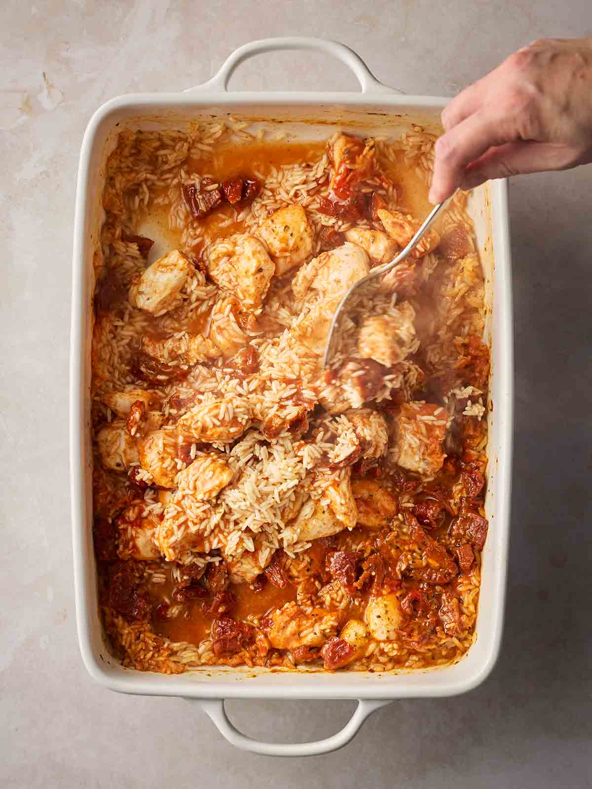 A hand with spoon stirring an oven dish fulled with chicken, rice and stock.