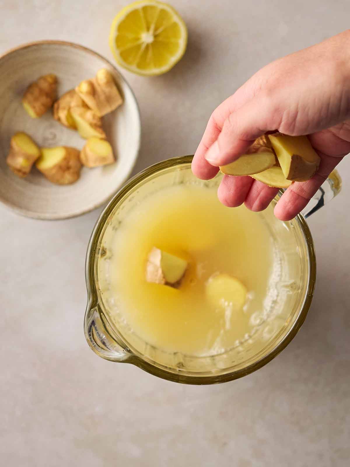 A jug of juice with fresh ginger in, a hand about to drop more into it, and a bowl underneath with more ginger and a sliced lemon to the side.