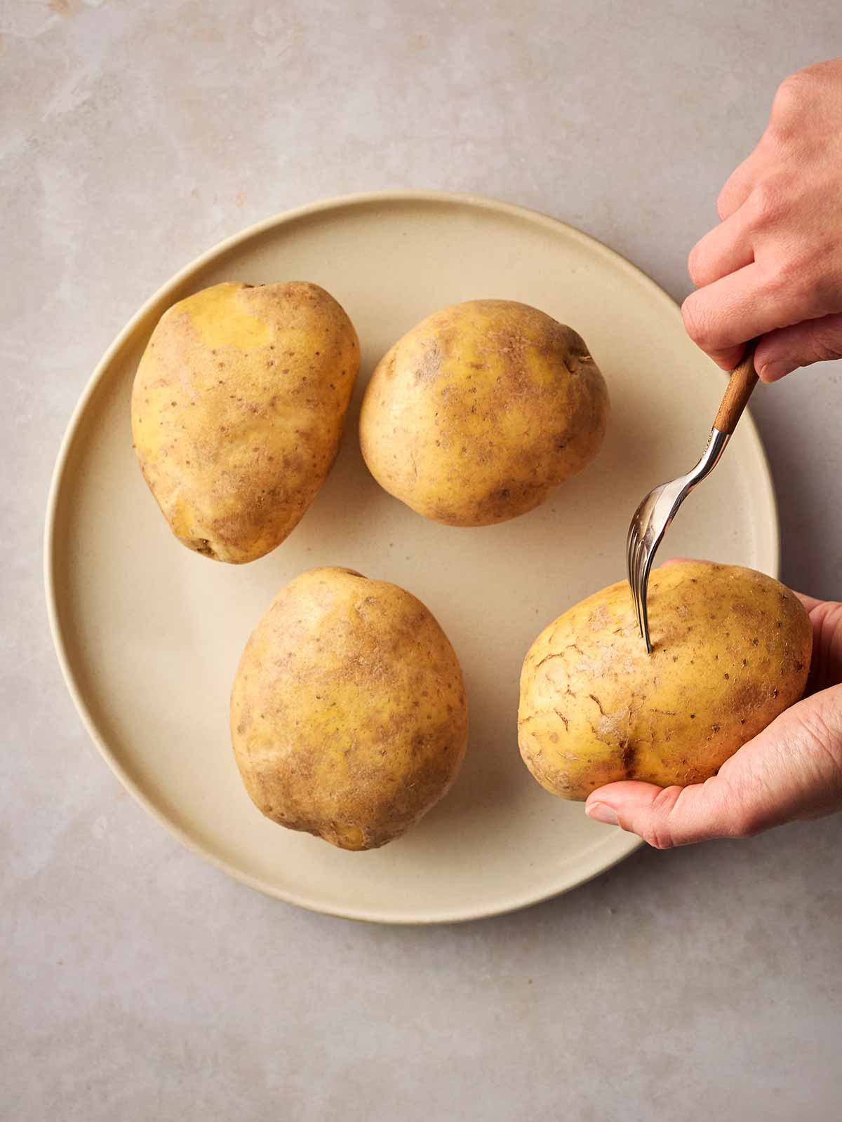 Two hands over a plate of potatoes, with a fork piercing one of them.