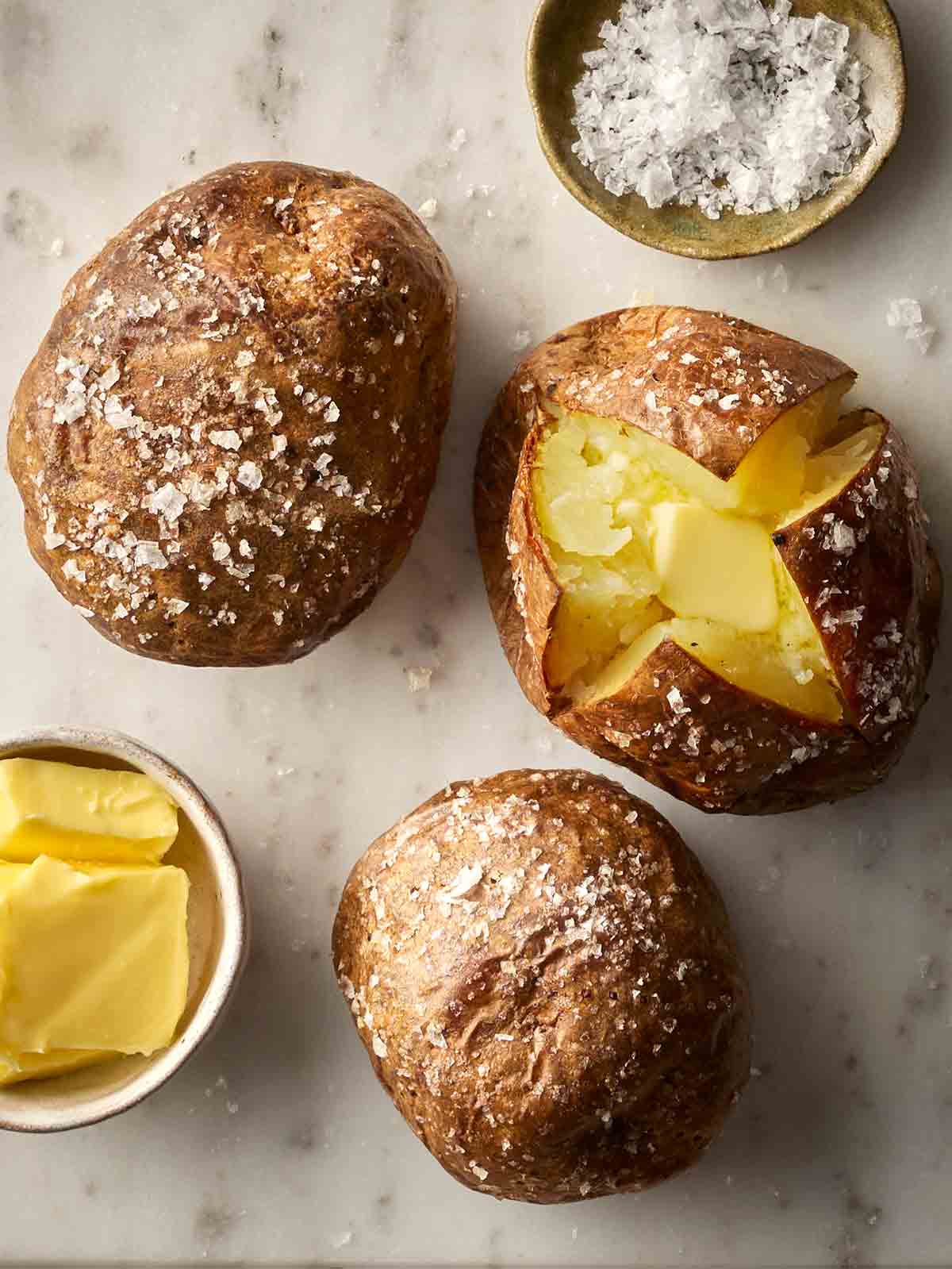Three perfectly cooked jacket potatoes on white surface, one of them cut with butter in the middle, two with salt over the top.