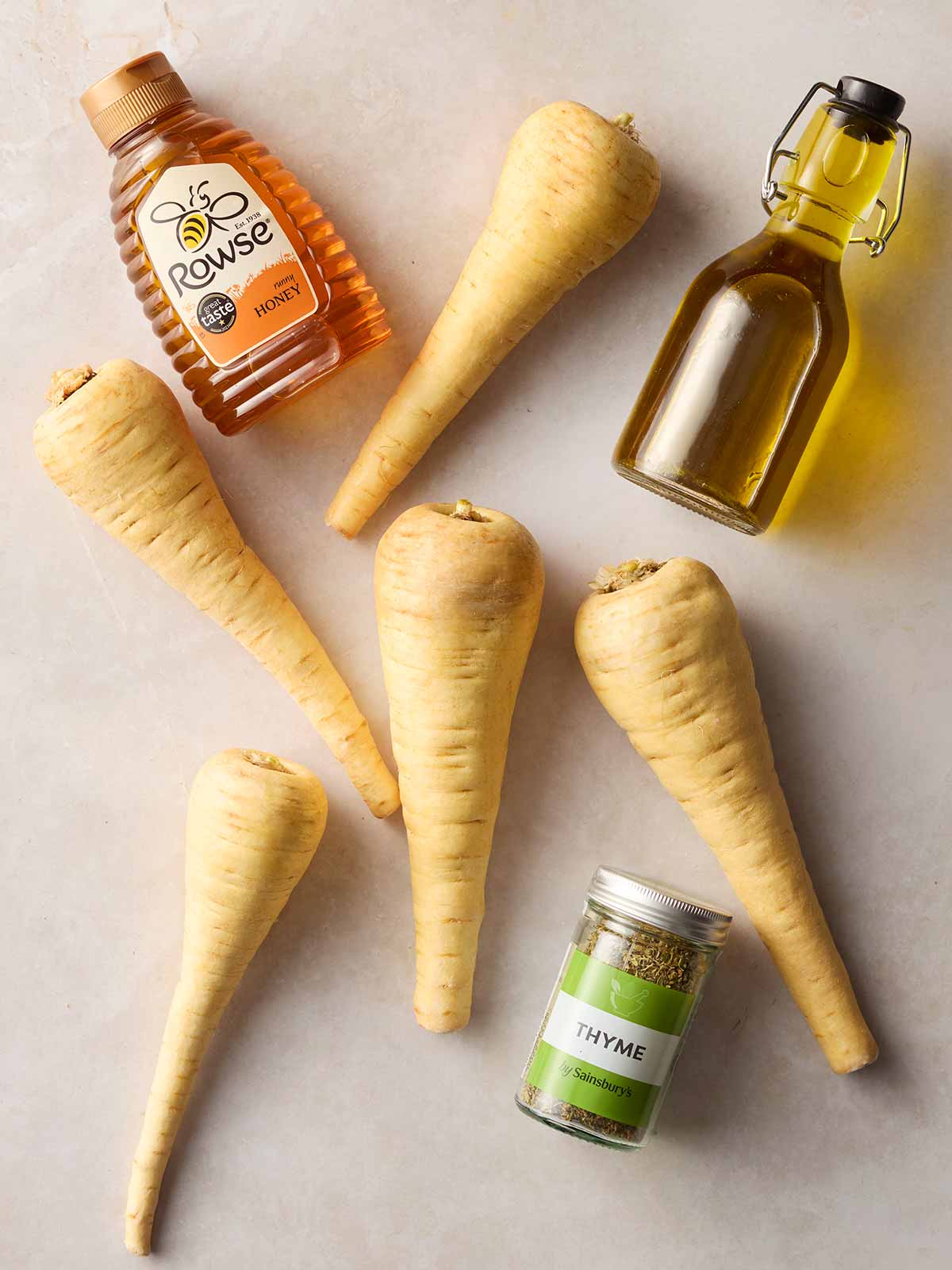 A white surface with parsnips, oil, honey and thyme laying on it.