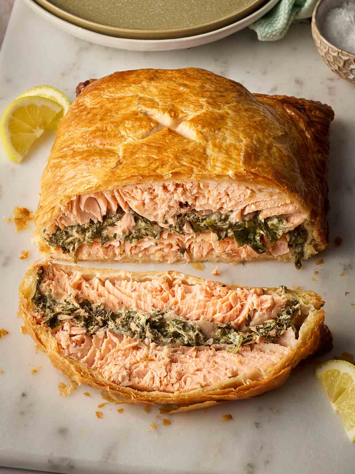 A sliced parcel of salmon in pastry with a spinach and cheesy filling, on a board ready to serve, with lemon slices around the side.