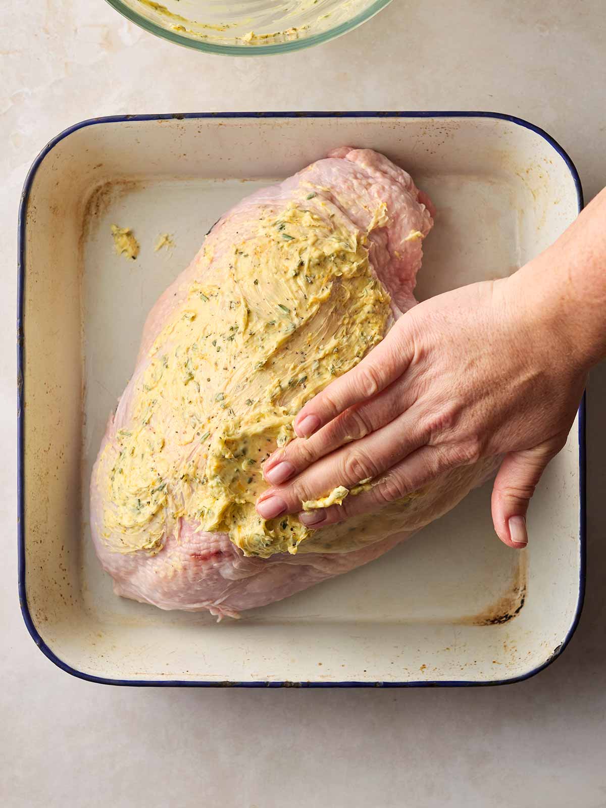 A hand smothering butter all over a raw turkey crown in an oven dish, getting it ready to roast.