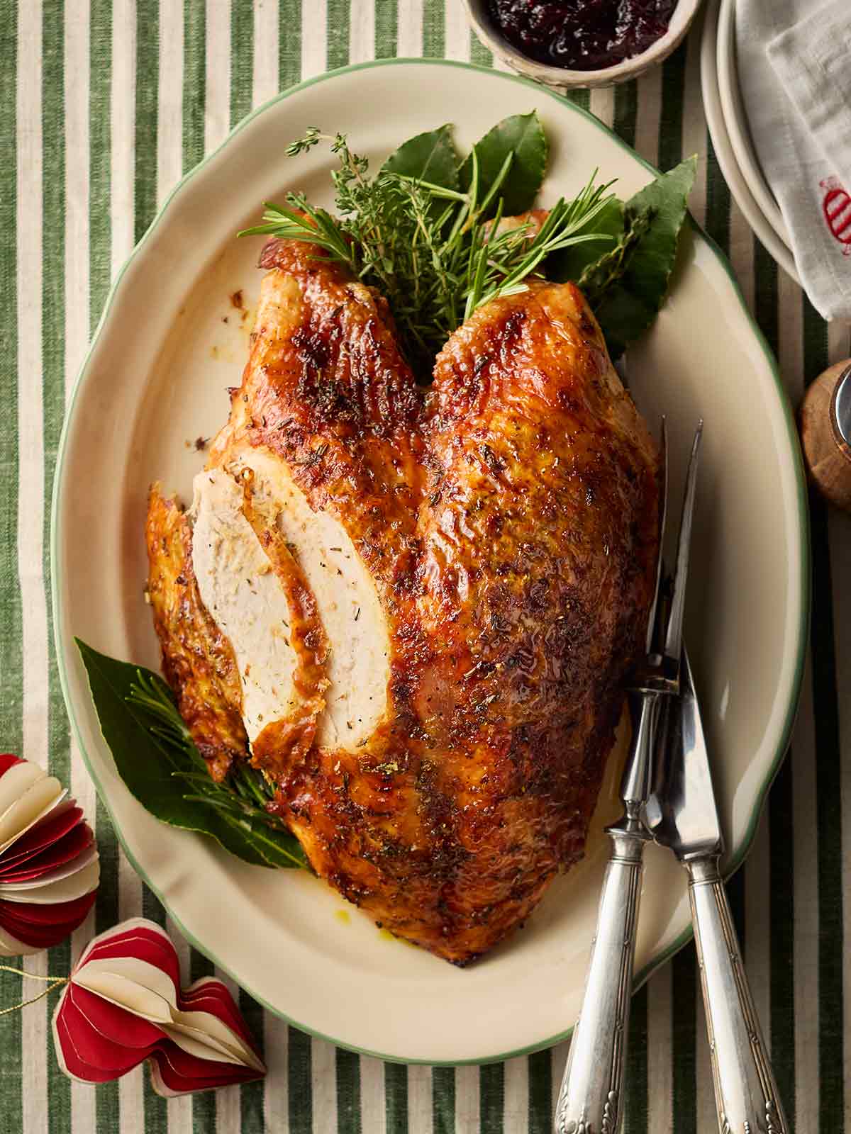 A roast turkey crown ready to serve from a platter with greens and herbs, a knife and serving fork, on a green and white tablecloth.