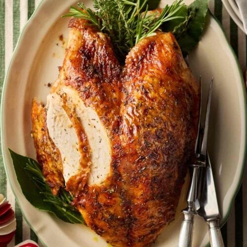 A platter with a cooked turkey crown and herbs, on a table with a green and white stripy tablecloth and Christmas decorations.