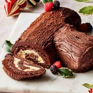 A Christmas yule log on a board, with a slice out to show the insides, and decorated with holly and berries.