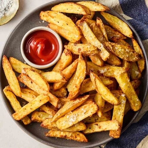 A plate of homemade chips with a side of ketchup on a surface with a blue and white tea towel and salt and pepper.