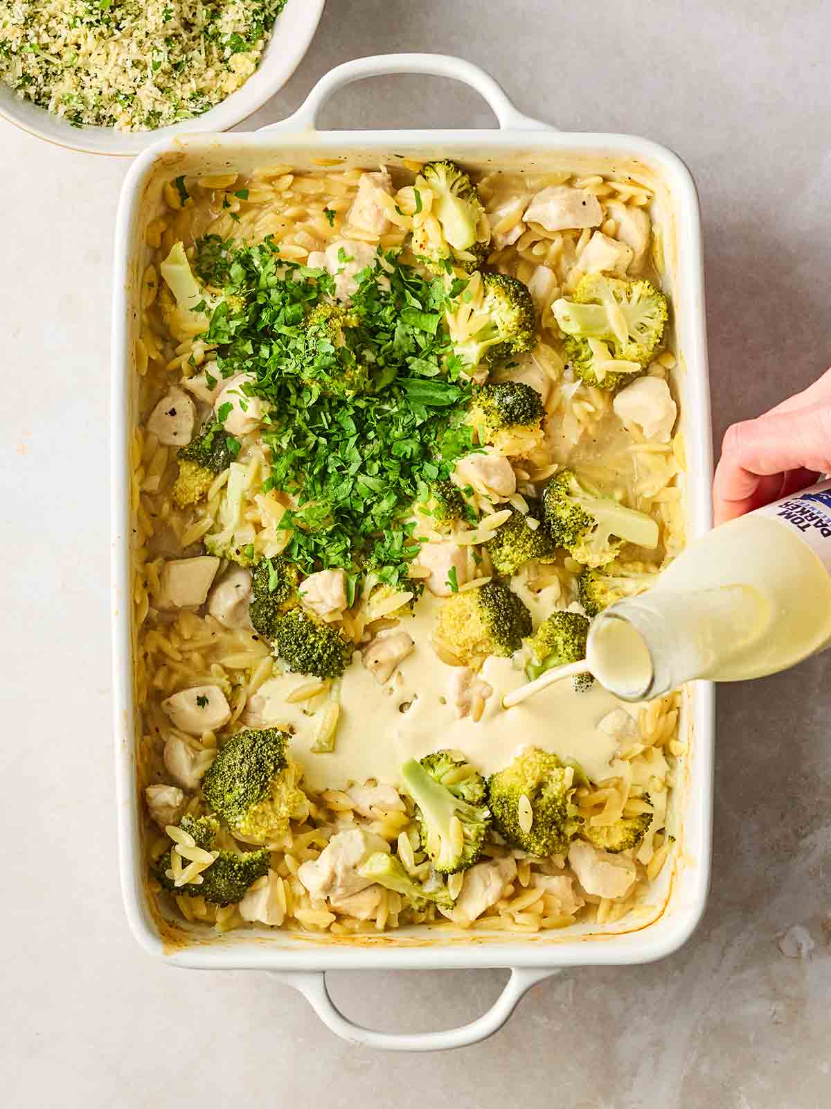 An oven dish filled with chicken, orzo pasta, broccoli, with a hand pouring in some double cream.