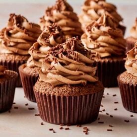 A batch of chocolate cupcakes with swirled chocolate icing and chocolate sprinkles, laid out on a table from the side.