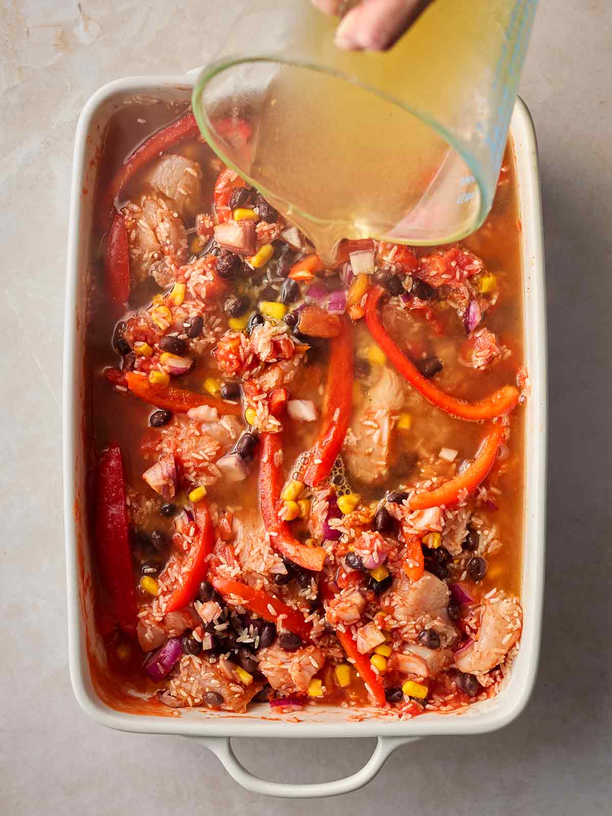 A baking dish with lots of raw ingredients inside to cook together, including chicken and rice, with a hand pouring stocking in.