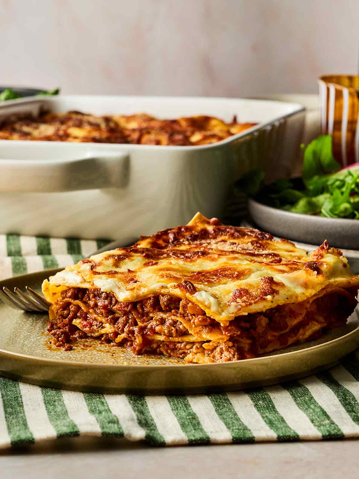 A plate in the foreground with a portion of lasagne on, in the background the oven dish with the rest of the lasagne, on a dining table.