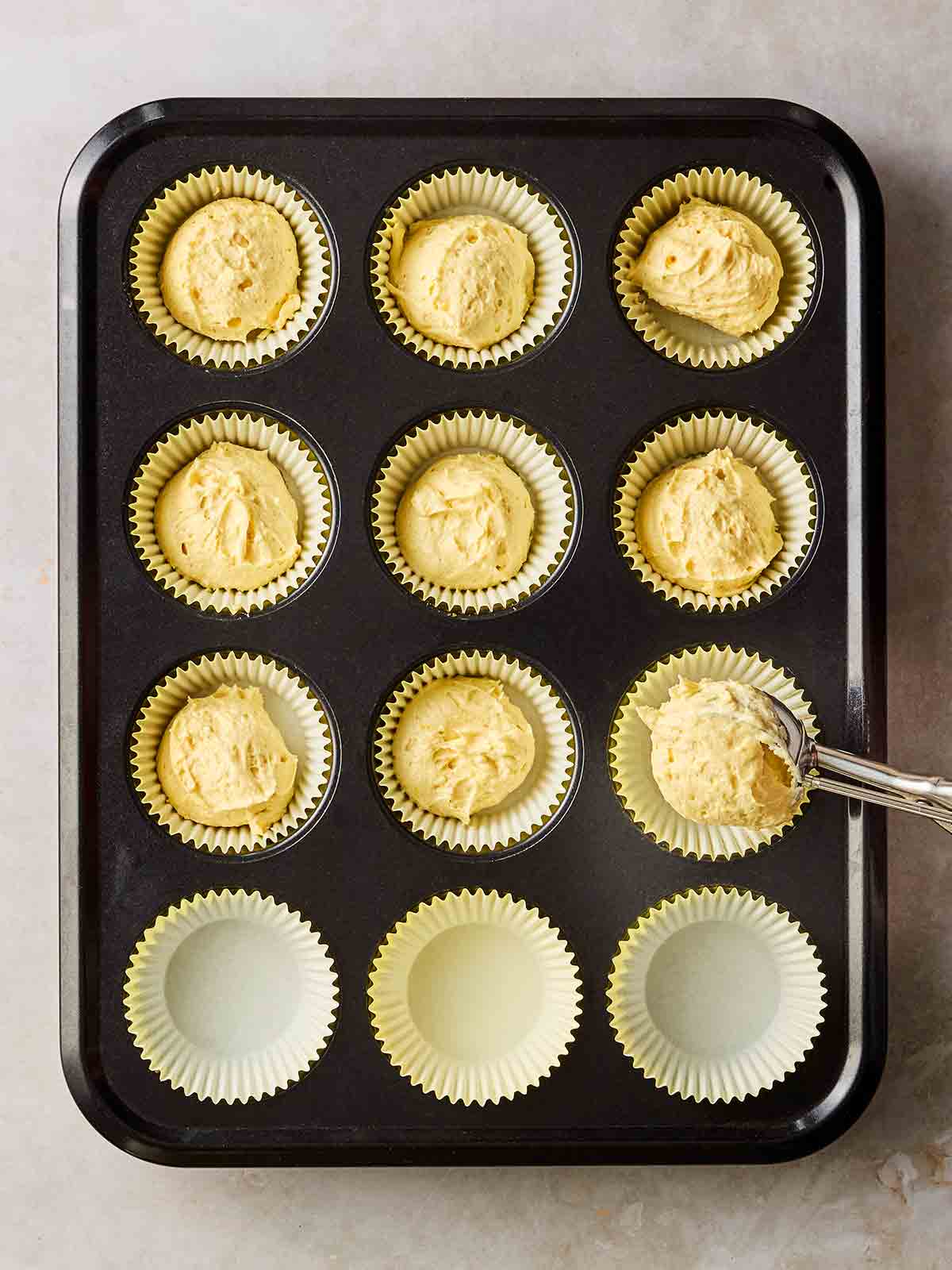 Cupcake batter in individual cases in a tray, with an ice cream scoop adding more.