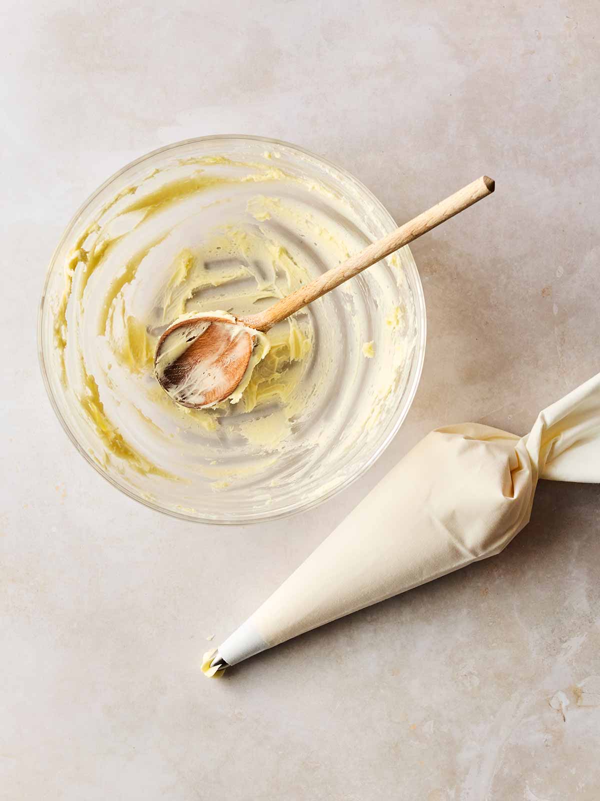 A glass bowl with a spoon in with cupcake mixture in a piping bag to the side.