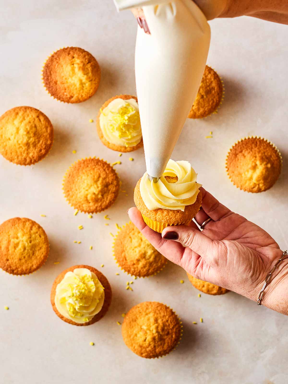 Two hands piping buttercream onto cupcakes, with more in the background.