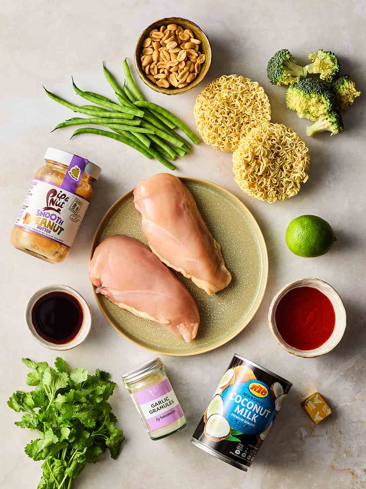 Ingredients laid out on a white surface, including raw chicken breasts, noodles, vegetables, coconut milk, peanut butter and more.