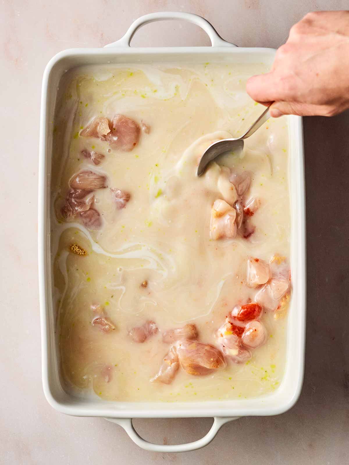 An oven dish filled with raw chicken breast chunks, coconut milk, spices and a hand is stirring the ingredients with a spoon.