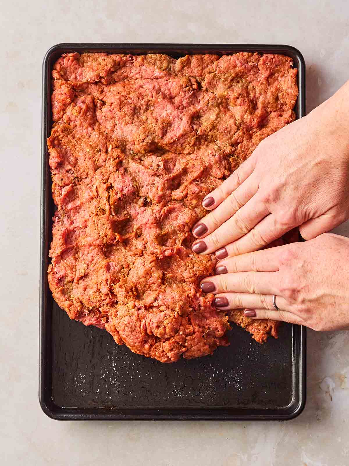 Two hands pressing a beef mince mixture into an oven dish for a meat sheet kebab recipe.