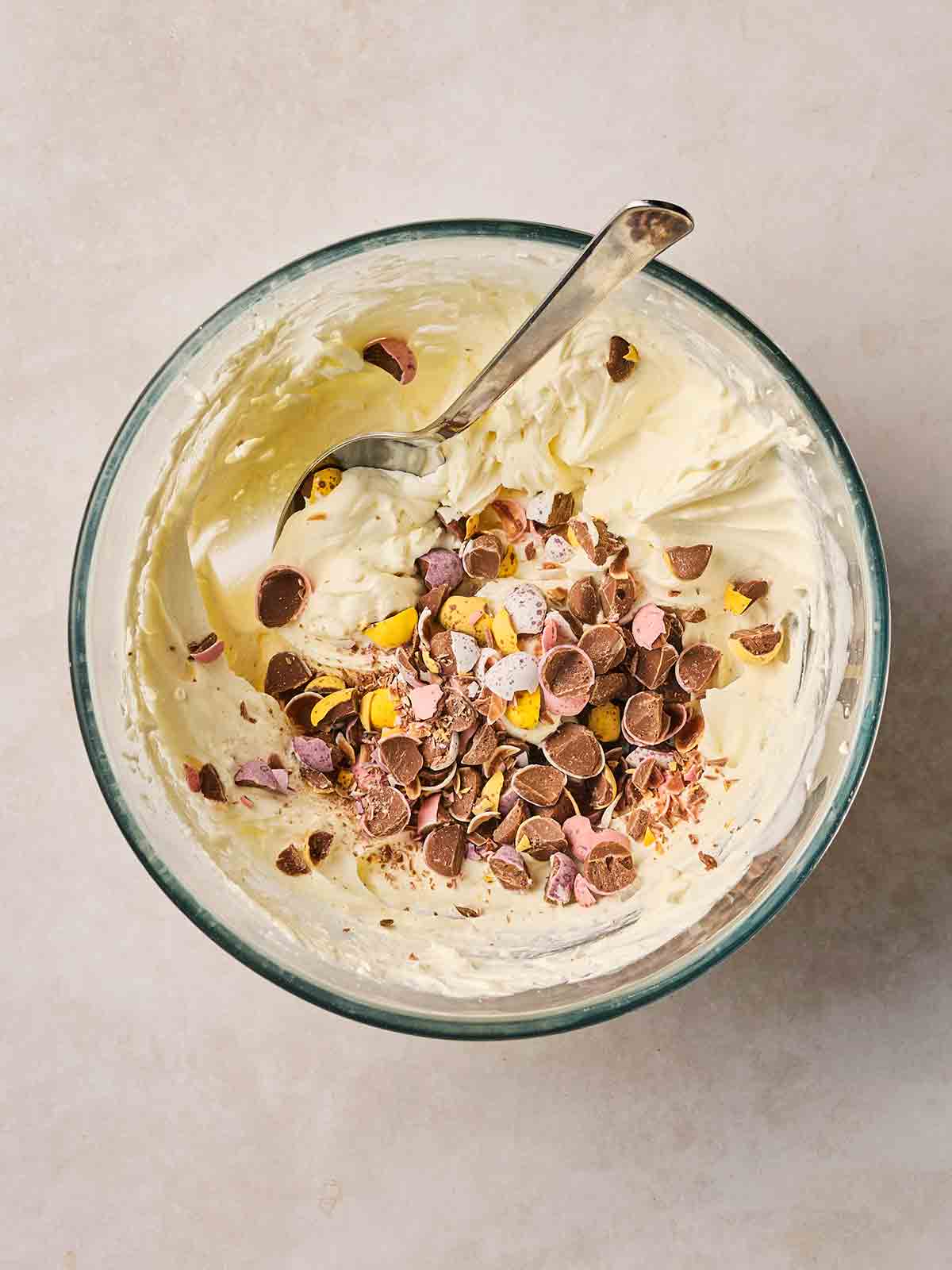 A glass bowl filled with whipped cream and a spoon stirring in crushed up mini eggs.