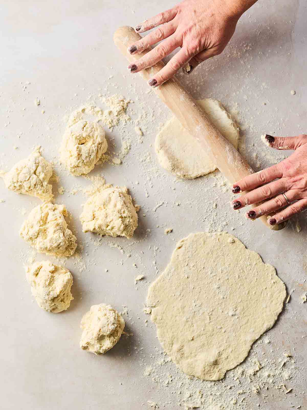 Portioned out flatbread dough on a white surface, being rolled out with a rolling pin.