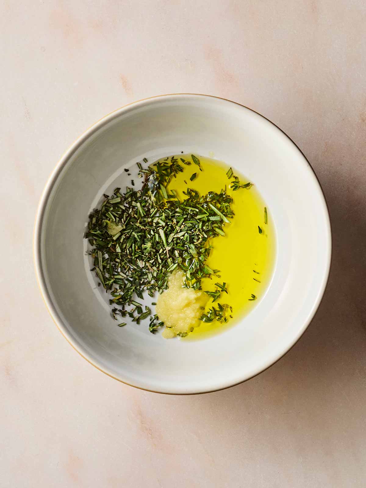 A white bowl with ingredients, unmixed, to make a marinade, including oil, garlic and chopped rosemary and thyme.