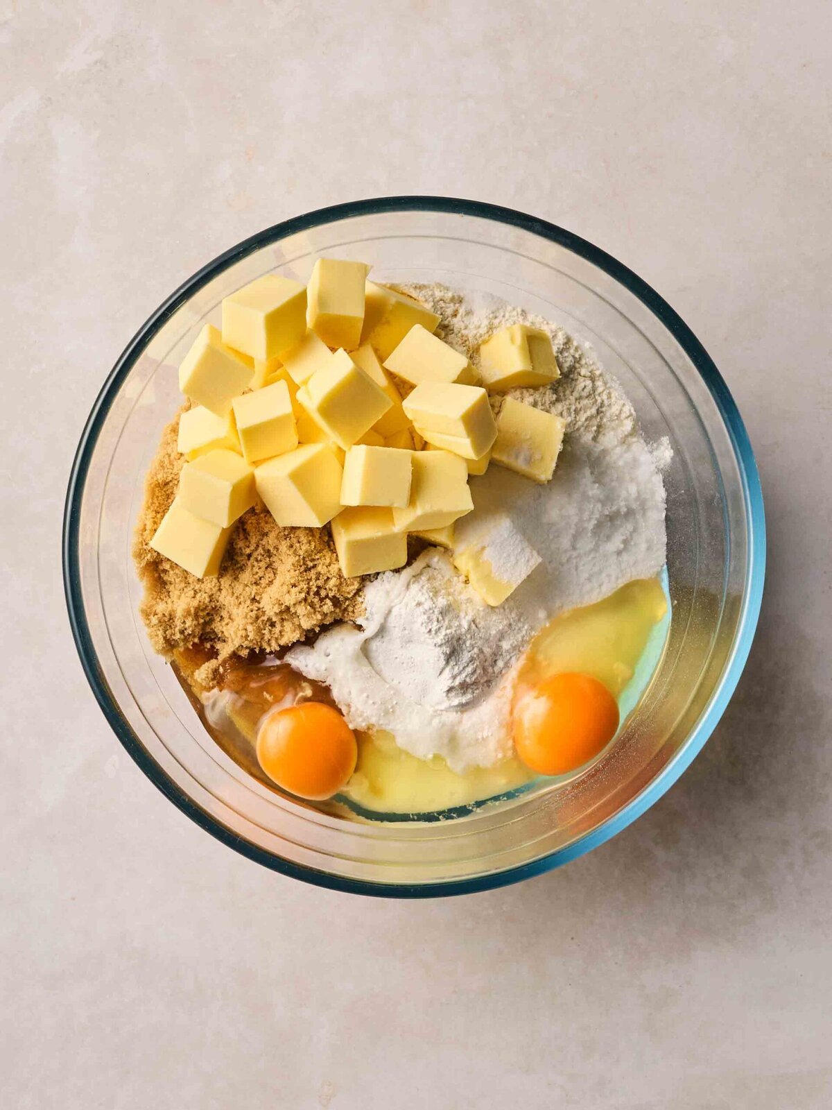 A glass bowl with unmixed ingredients of cubed butter, sugar, flour and eggs.