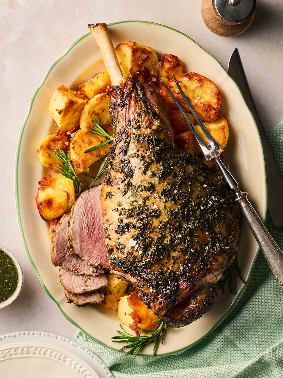 A platter with a roasted leg of lamb on top, with roast potatoes around the edges, and a few sliced pieces. A fork ready to slice to the side.