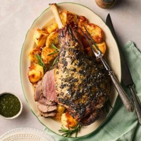 A roast leg of lamb on a platter with roast potatoes on the side, with a few slices ready to eat, mint sauce on the side, and a carving knife and fork ready to slice up the rest.